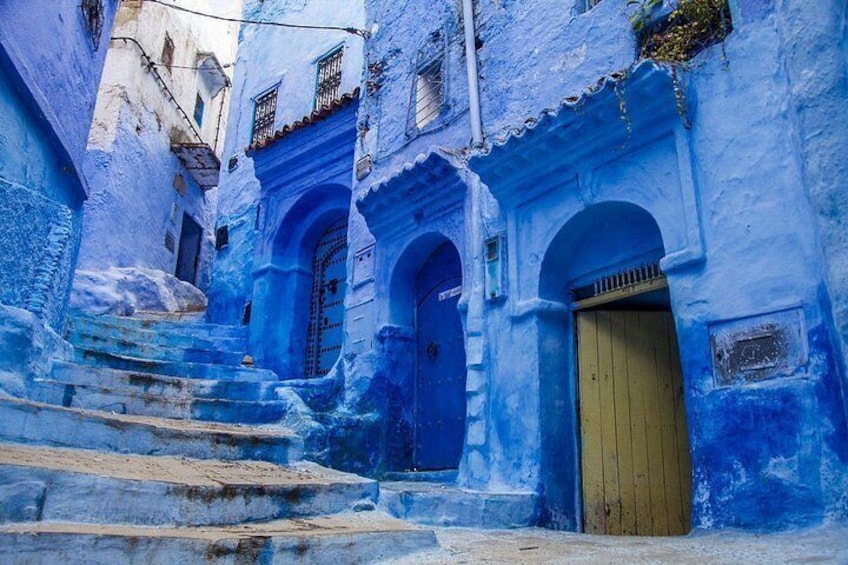 Une ruelle étroite bordée de bâtiments peints en bleu, avec une porte en bois et des marches en pierre. 1