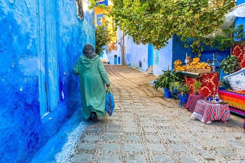 Una calle estrecha con una persona caminando, paredes azules y un puesto de frutas. 3