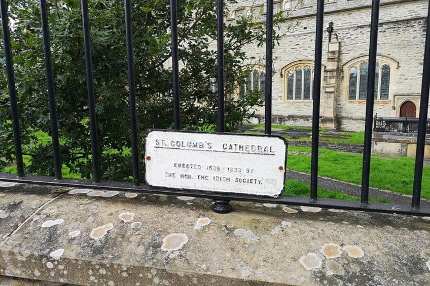 A signboard with the text 'St. Columba's Cathedral' and 'Erected 1828 by the Hon. the Irish Society' in front of a stone wall with a metal fence. 5