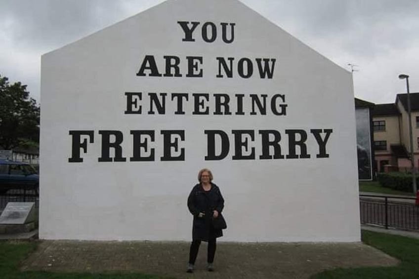 A person standing in front of a large white sign with black lettering that reads 'YOU ARE NOW ENTERING FREE DERRY.' 1