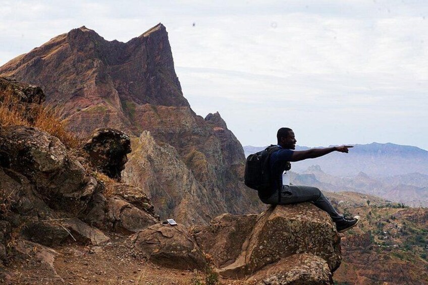 Una persona sentada sobre una roca con una mochila, señalando hacia una cordillera. 3