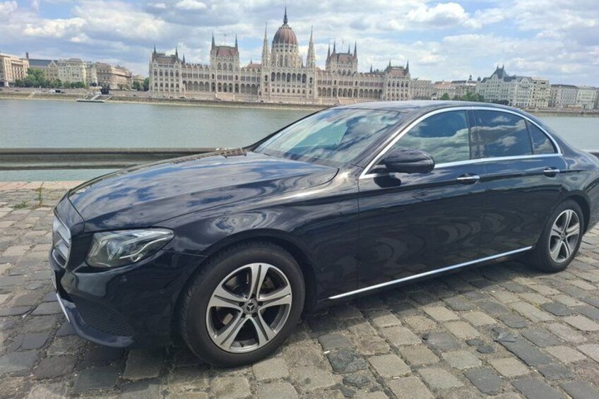 Eine schwarze Limousine parkt auf Kopfsteinpflaster, im Hintergrund fließt ein Fluss und steht ein historisches Gebäude. 5
