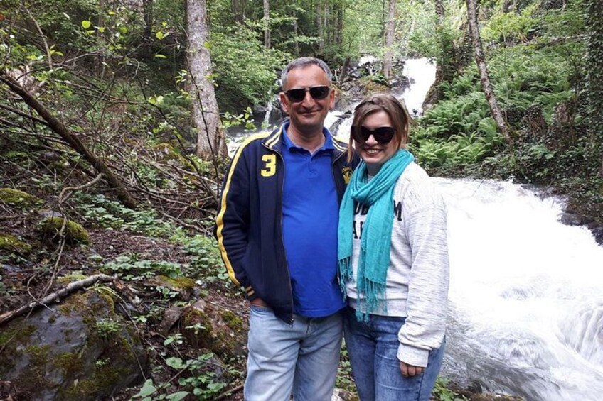 Two people standing by a flowing river in a forested area. 5