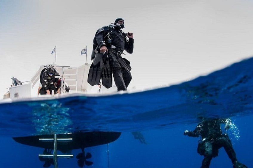 Ein Taucher in schwarzer Ausrüstung auf einem Boot, ein weiterer Taucher unter Wasser und ein dritter Taucher in anderer Position untergetaucht. 1