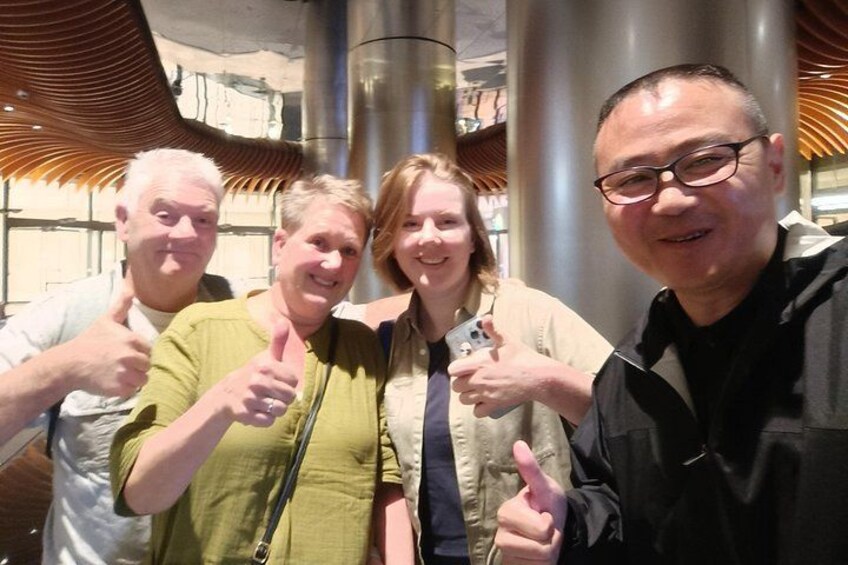 Four people giving thumbs up in front of a modern architectural structure with curved metal elements. 4