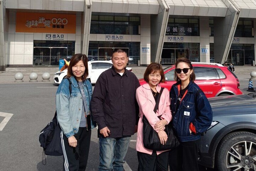 Four people standing in front of a building with signage, a red car, and a white car parked nearby. 3