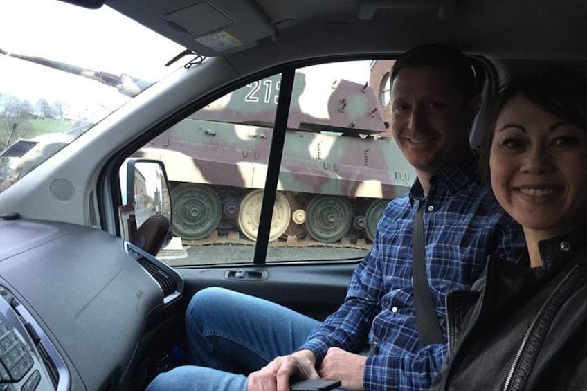 A man and a woman in a car with a military vehicle in the background. 1