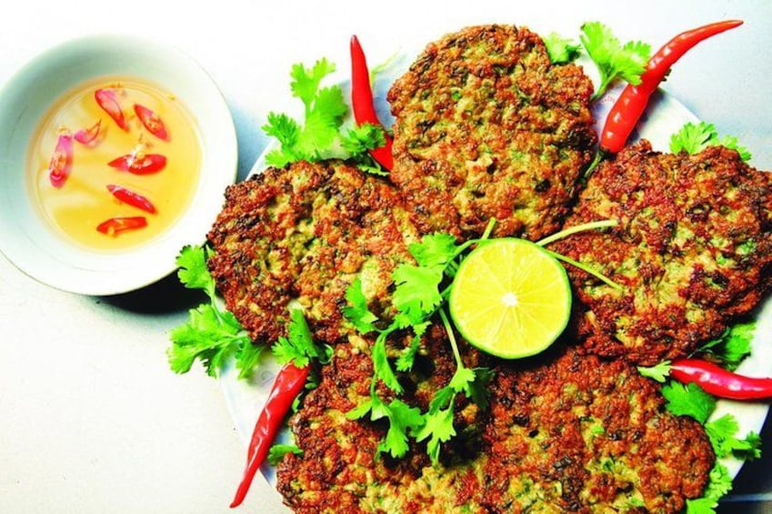 A plate of fried patties garnished with cilantro and red chili peppers, accompanied by a bowl of sauce and a slice of lime. 2