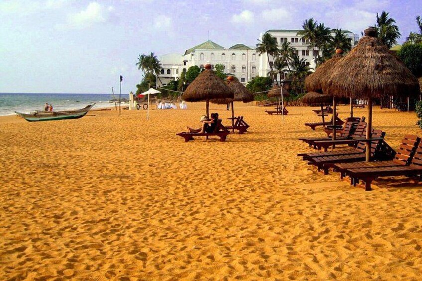 Een zanderig strand met rieten parasols, ligstoelen en een gebouw op de achtergrond. 1