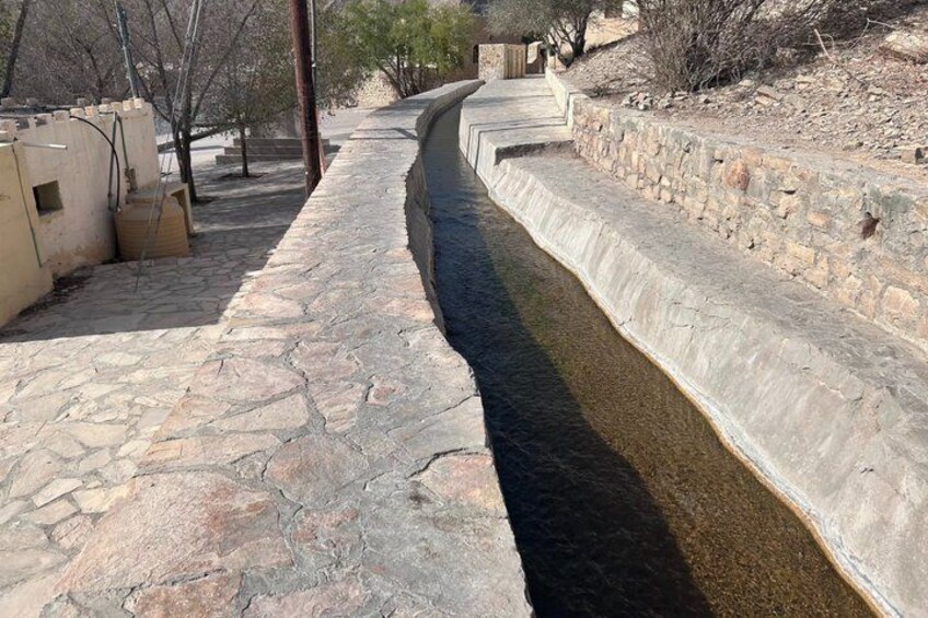 Ein mit Stein ausgekleideter Kanal mit Betonrand, umgeben von trockener Vegetation und einer Steinmauer. 5