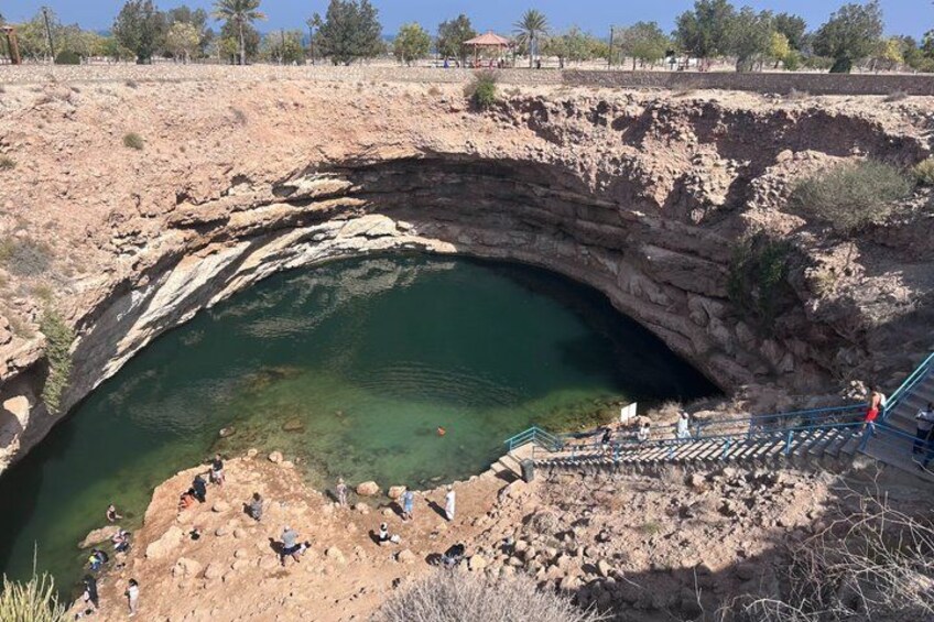 A natural rock formation with a water-filled basin, a staircase leading down, and people around the area. 2