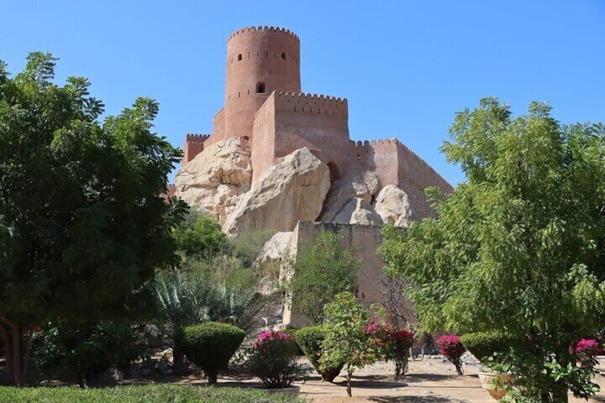 A historic castle with a round tower, set against a backdrop of lush greenery and flowering plants. 1