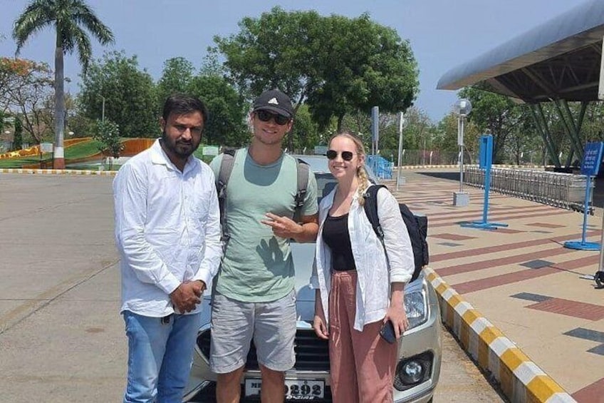 Three people standing in front of a car, a paved area with trees, and a covered structure in the background. 3