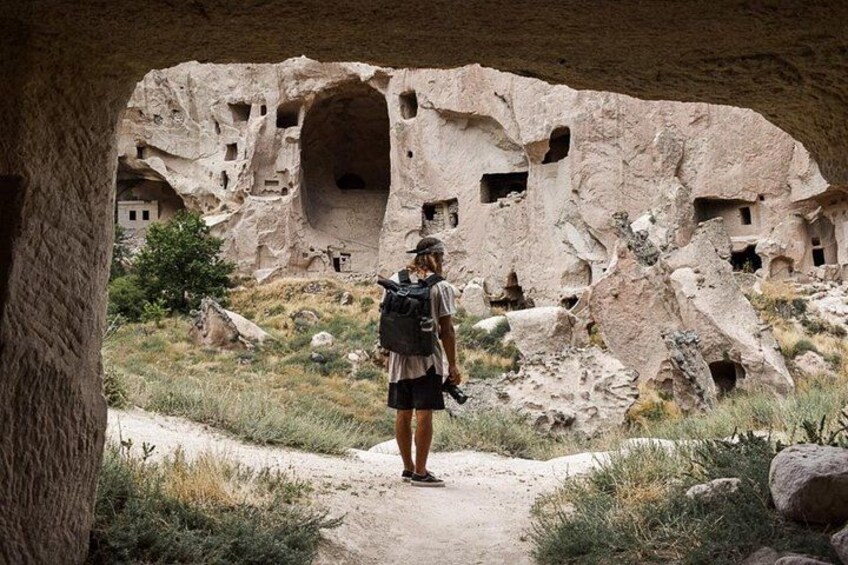 A person standing on a dirt path with a backpack, looking at ancient rock structures with multiple openings. 2