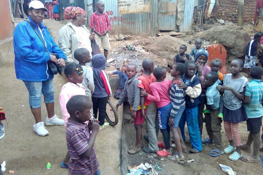 A group of children gathered around a man in a blue jacket, standing on a dirt ground with a makeshift shelter in the background. 1