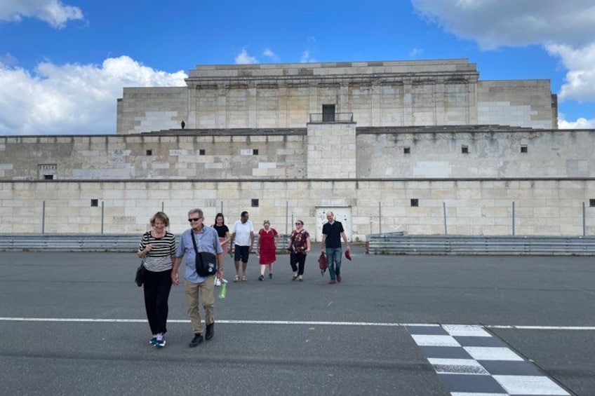 A group of people walking in front of a large stone building with a checkered pattern on the ground. 3