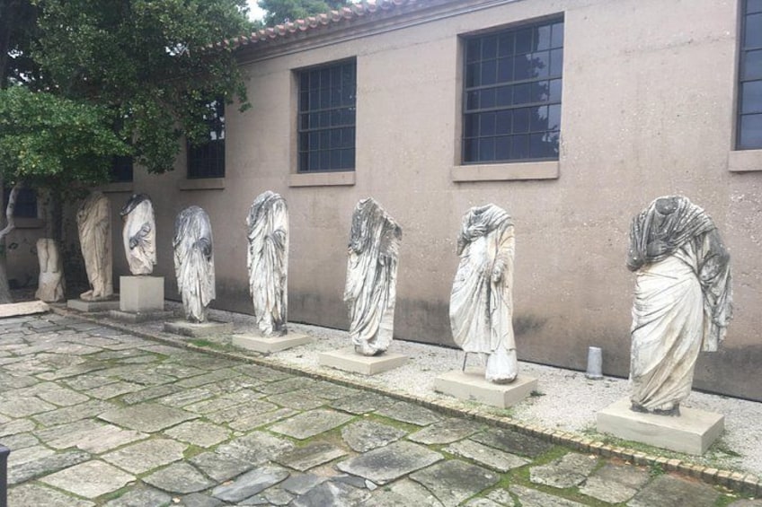 Eine Reihe klassischer Marmorstatuen in einem Innenhof mit Steinpflasterung und einem Gebäude im Hintergrund. 4
