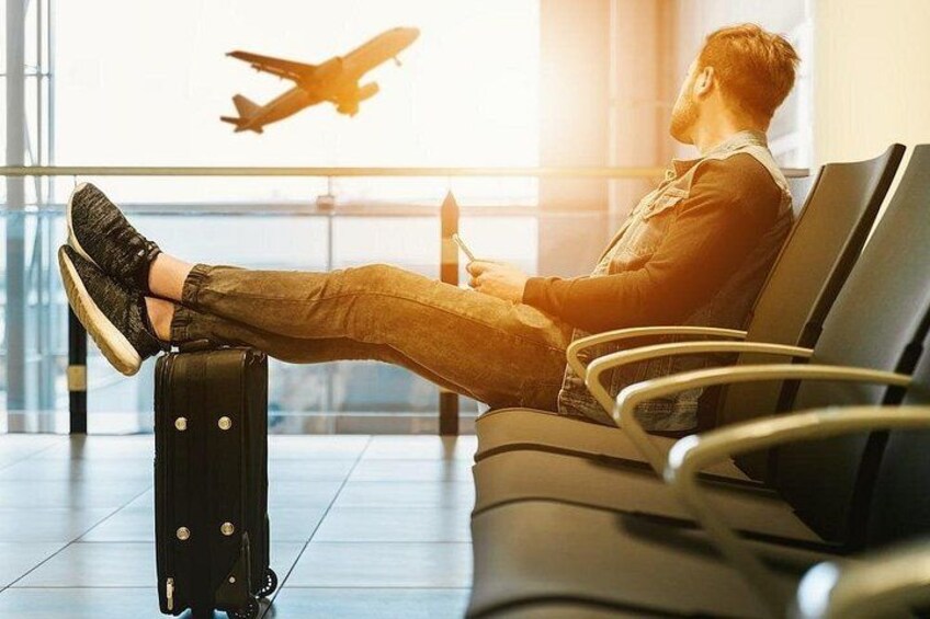 Una persona sentada en un banco del aeropuerto con una maleta, un avión al fondo y una ventana con luz natural. 1