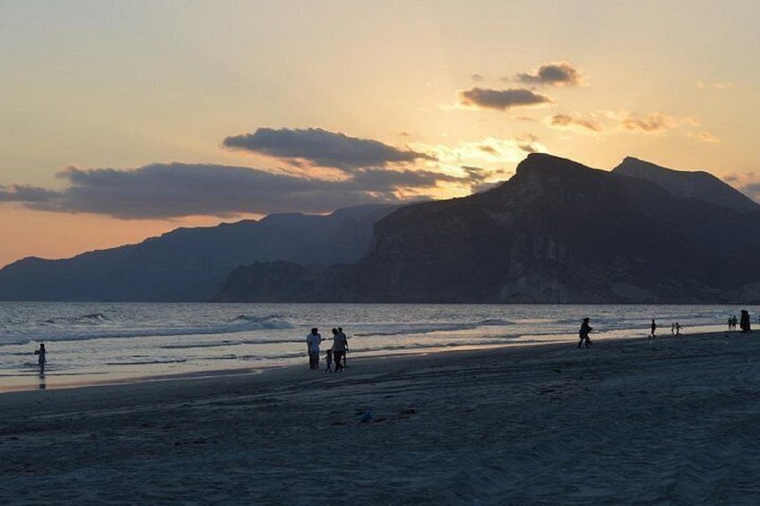 A beach with people walking and playing, mountains in the background, and a sunset. 2
