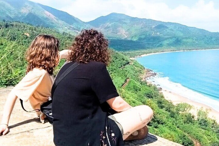 Two people sitting on a bench overlooking a coastal landscape with mountains in the background. 1