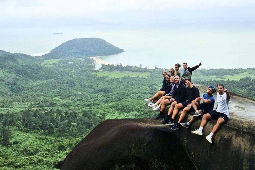 A group of people sitting on a rock overlooking a lush landscape with a body of water and a hill in the distance. 4