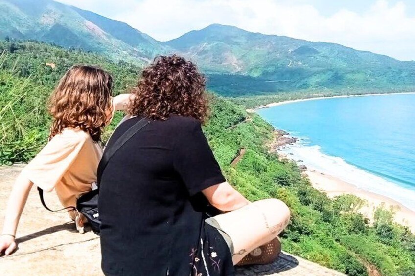 Two people sitting on a ledge overlooking a coastal landscape with mountains in the background. 3