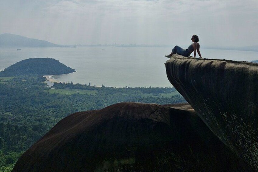 A person sitting on a cliff overlooking a coastal landscape with a body of water and distant hills. 1