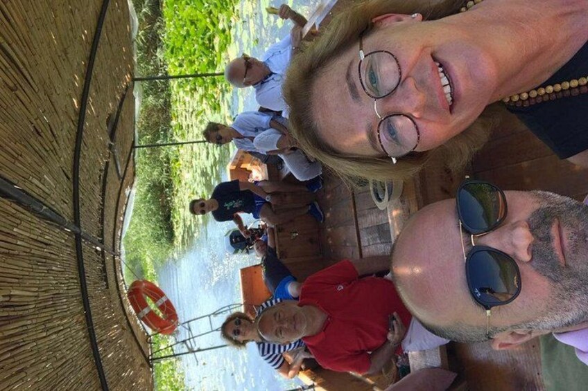 A group of people standing on a wooden deck by a river, with a bridge in the background. 1