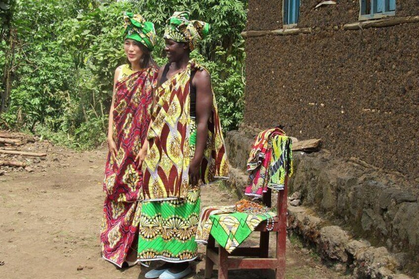 Two people in colorful traditional attire standing in front of a rustic building with a thatched roof. 3