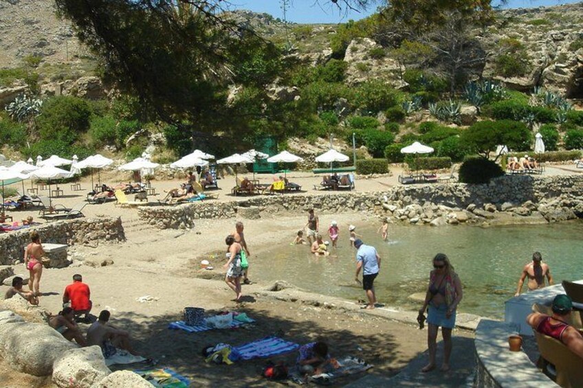 Ein Strand mit Personen, die sonnenbaden, schwimmen und spazieren gehen. Sonnenschirme und Liegestühle sind vorhanden. 2