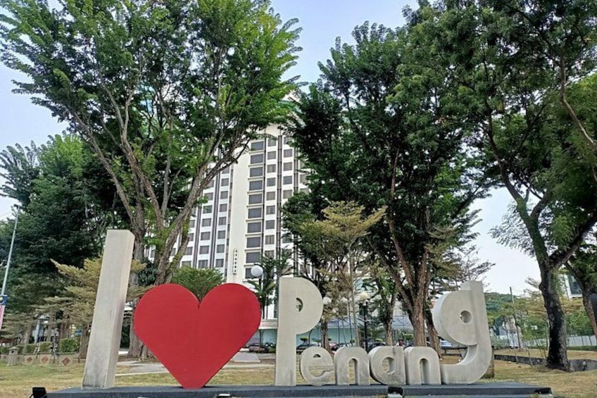 Ein großes, rotes Herz und der Schriftzug 'I love Penang' in einem Park mit Bäumen und einem hohen Gebäude im Hintergrund. 1