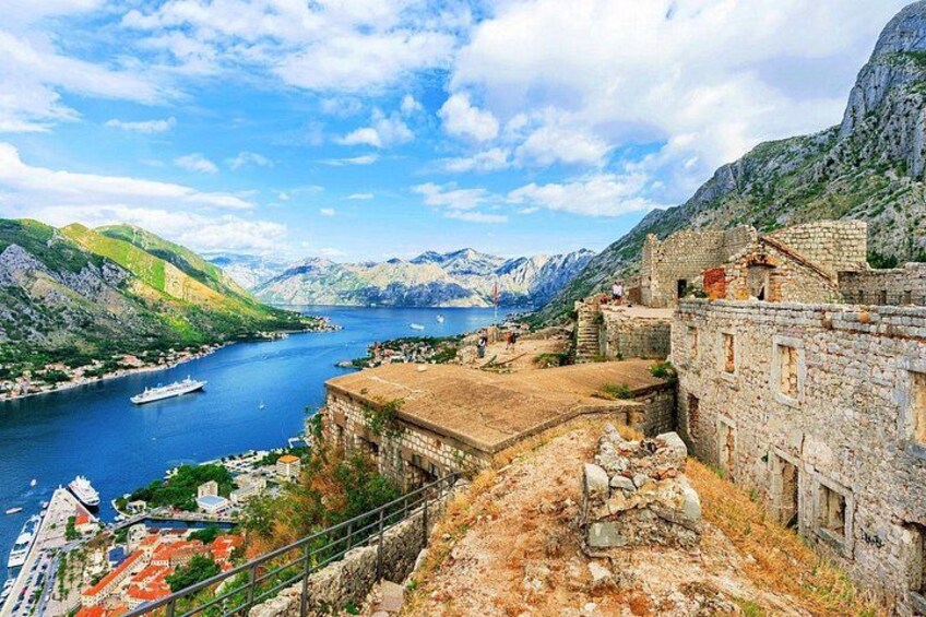 A coastal town with historic stone buildings, a harbor with boats, and mountainous terrain in the background. 1