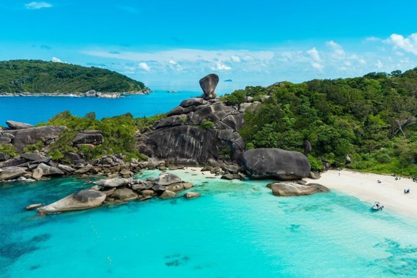 Una spiaggia con grandi massi, acqua turchese cristallina e una collina boscosa sullo sfondo. 2