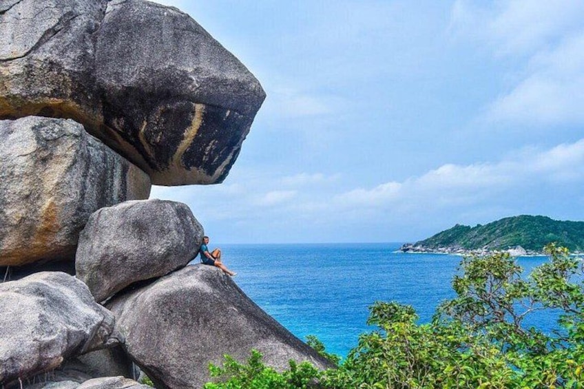 Una persona seduta su una grande formazione rocciosa affacciata sul mare, con una piccola isola sullo sfondo. 3