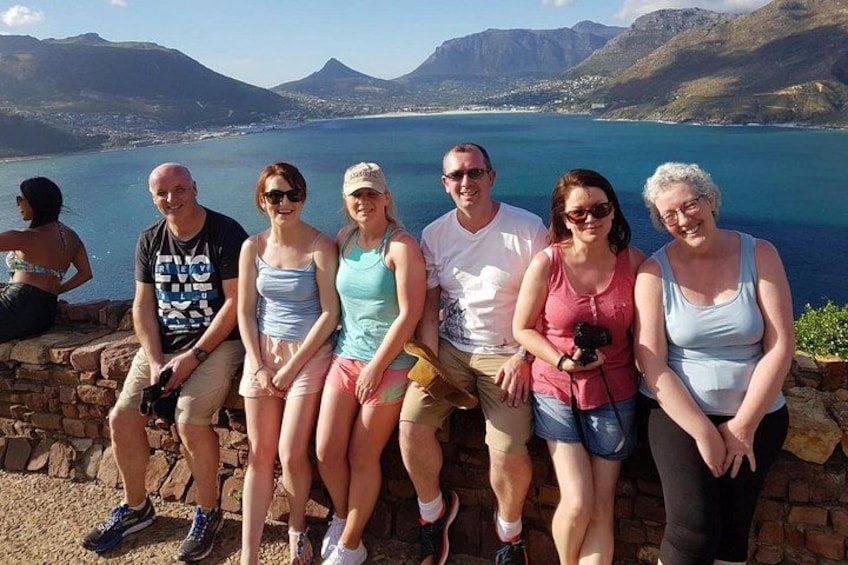 Un gruppo di persone sedute su un muro di pietra con vista su un lago e montagne sullo sfondo. 5