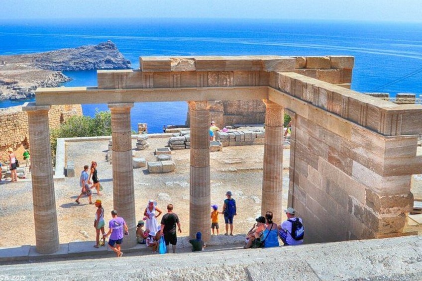 Ancient ruins with columns, people walking, and the sea in the background. 3