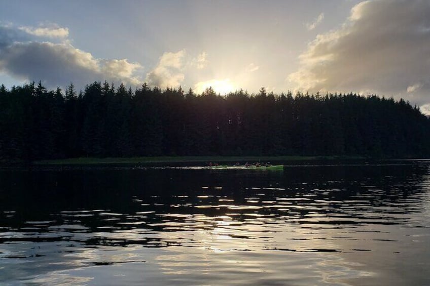 Et roligt sø med en grøn kajak i det fjerne, omgivet af en tæt skov og solnedgang på horisonten. 3