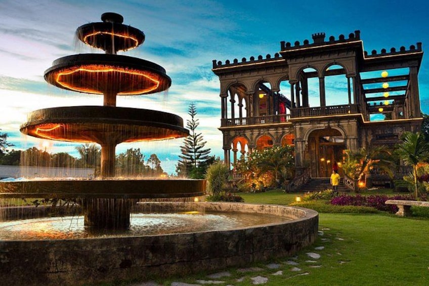 Une fontaine à plusieurs niveaux, dont l’eau jaillit en cascade, est entourée d’une pelouse bien entretenue et d’un bâtiment historique doté d’arcades et de balcons. 1