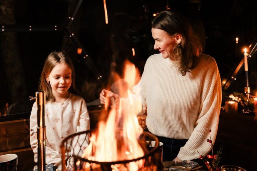 A person and a child near a campfire with a metal grill, candles, and a dark background. 1