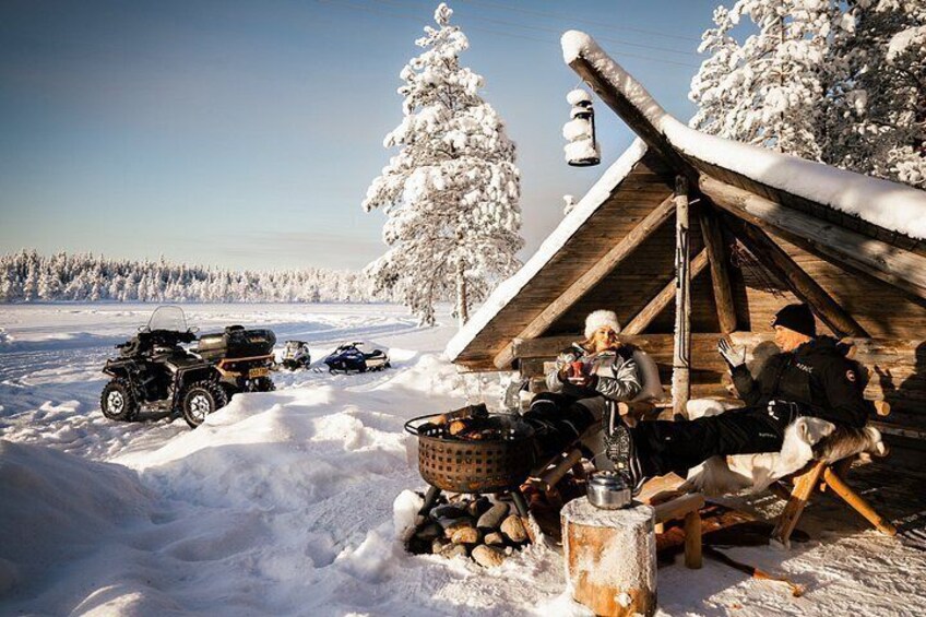 A snowy outdoor scene with a wooden cabin, ATVs, and people gathered around a fire pit. 2