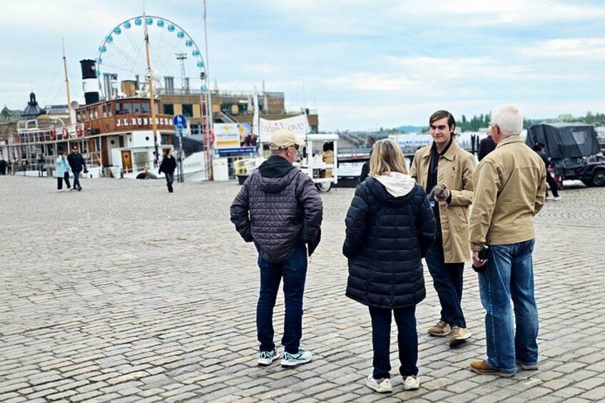 Eine Gruppe von Menschen steht auf einem Kopfsteinpflasterweg, im Hintergrund sieht man ein Riesenrad und ein Gebäude. 1