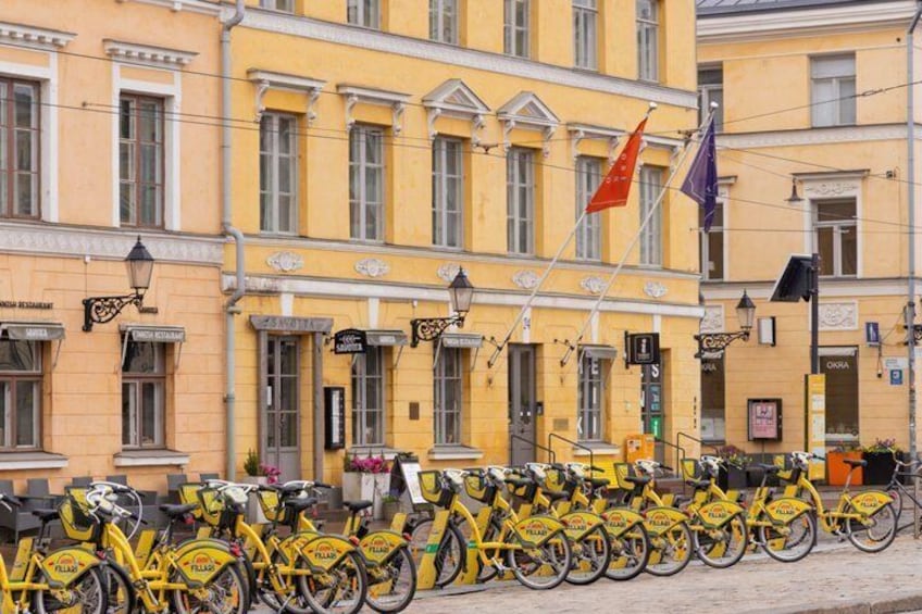 A row of yellow bicycles parked in front of a yellow building with flags and shop signs. 5