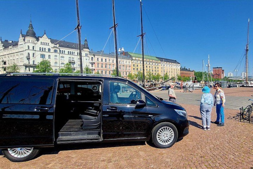 Una camioneta negra estacionada sobre adoquines, con un edificio histórico y altos barcos de vela al fondo. 3