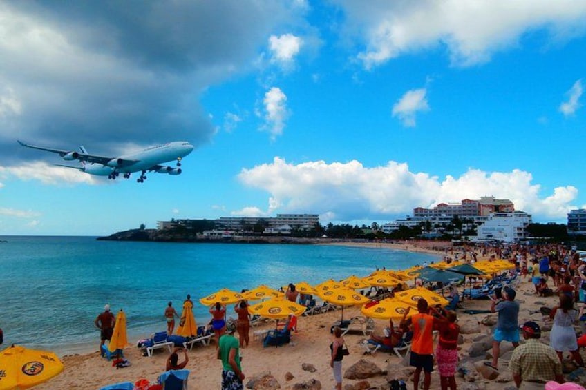 A beach with people, umbrellas, and a plane in the sky. 1