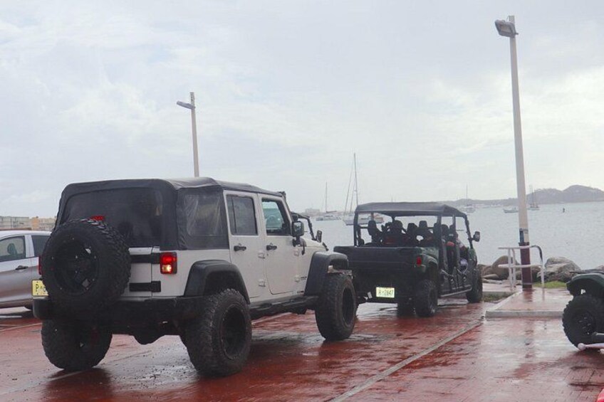 A white off-road vehicle parked on a wet surface near a body of water with boats in the background. 4