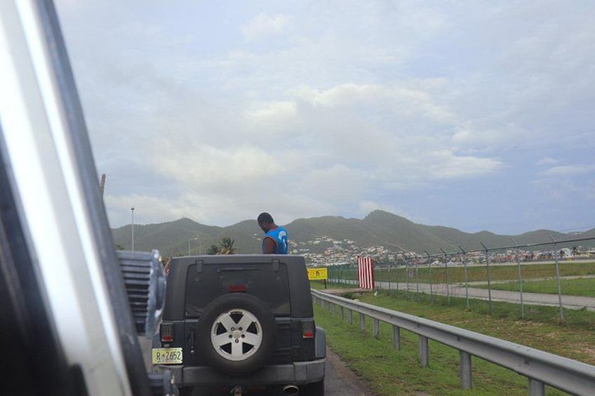 A vehicle on a road with a person standing on the back, a guardrail, and mountains in the background. 5