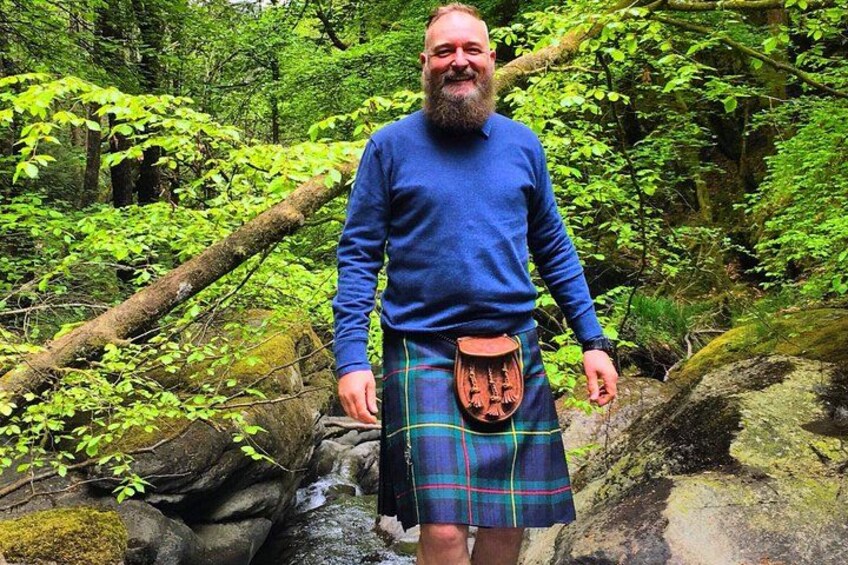 A person wearing a blue sweater and a tartan kilt standing in a forested area with a stream. 4
