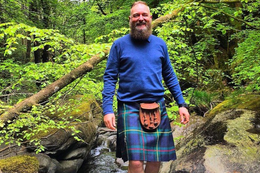 A person in a blue sweater and kilt standing in a forested area with a stream. 3