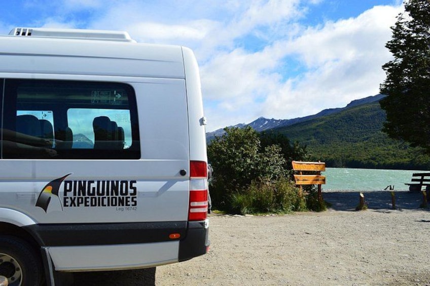 A white van with 'Pinguinos Expediciones' written on the side parked near a lake with mountains in the background. 2