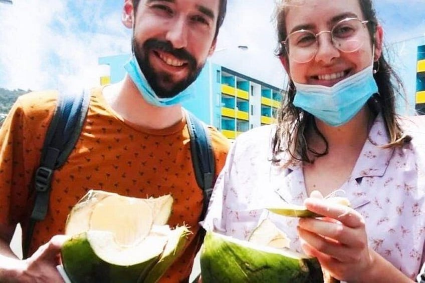 Two people holding coconuts, one with a backpack, and a building with blue and yellow stripes in the background. 2